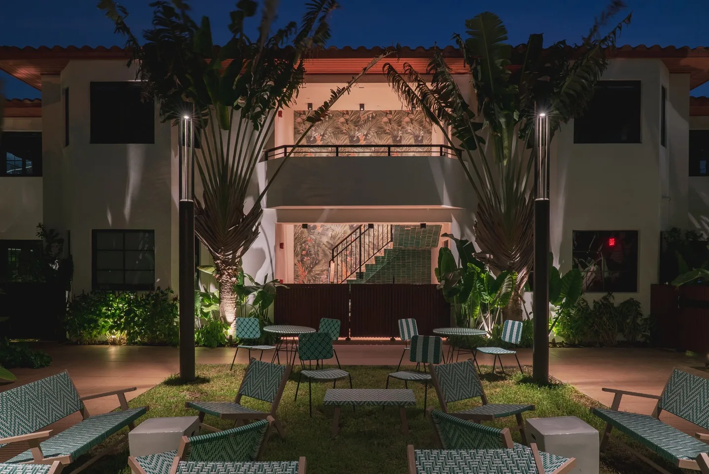 The lit house at evening over the courtyard's green chairs and tables.