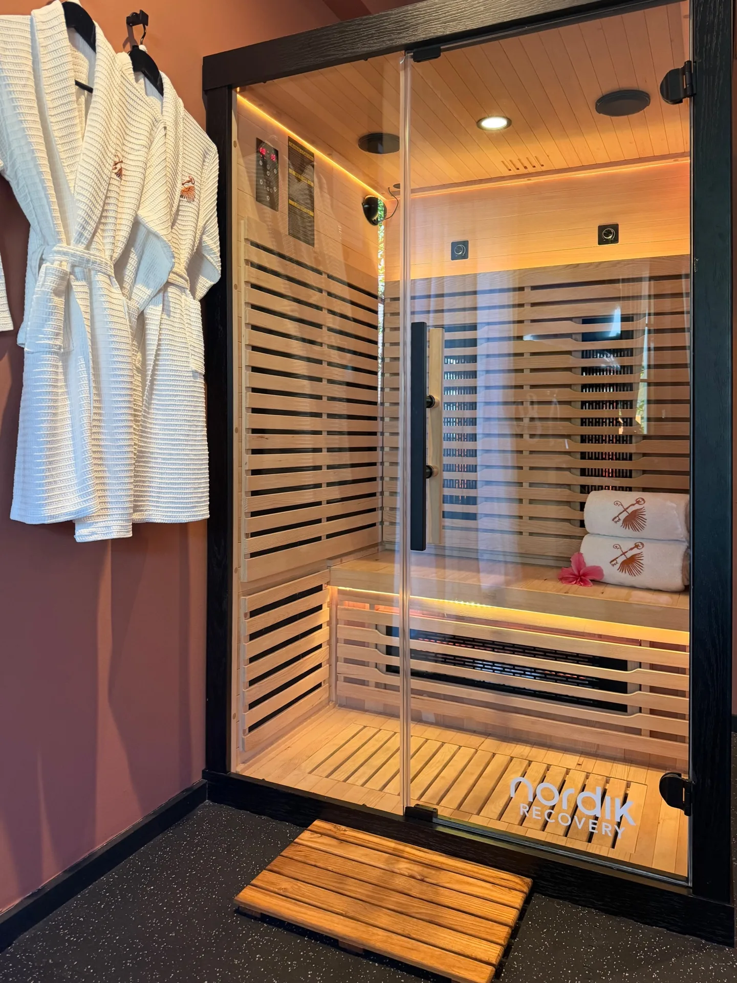 Infrared sauna cabin with a glass door and pale wood slats, two white robes hanging beside it on a terracotta wall