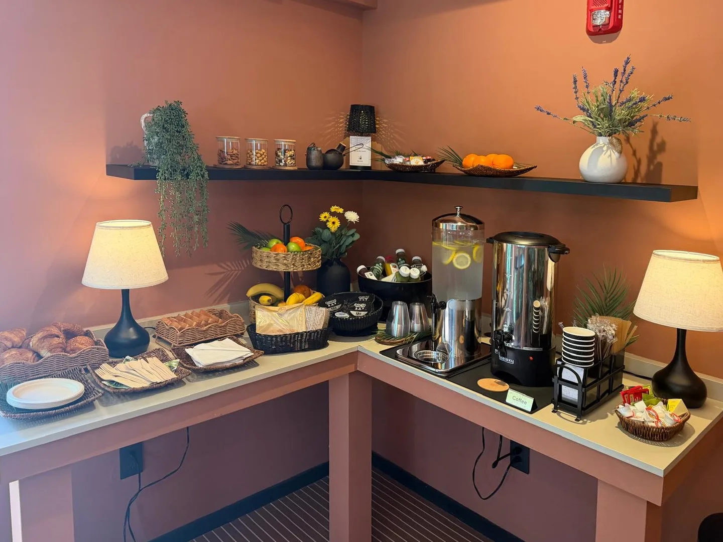 El rincón de desayuno en Maison Felix: croissants, bandejas de fruta, jugos sobre hielo, un dispensador de agua con limón y una cafetera en un mostrador de dos caras bajo una repisa oscura, lámparas encendidas contra paredes de terracota
